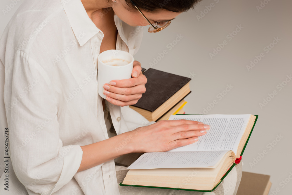 Wall mural young brunette woman in glasses reading book and holding cup of coffee isolated on grey