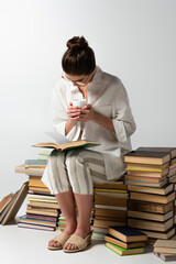 full length of young brunette woman in glasses sitting on pile of books while reading and holding cup on white