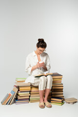 full length of young woman in glasses sitting on pile of books while reading and holding cup on white
