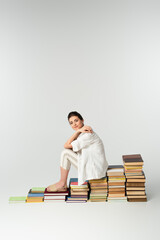 full length of pleased young woman in sandals sitting on pile of books on white