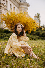 Young barefoot woman wearing a yellow muslin dress, sitting in a garden with green grass and...