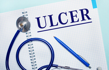 On a light blue background, an open notebook with the word ULCER, a blue pen and a stethoscope....