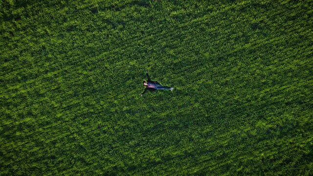 Aerial view of young slim woman lying motionless in large green field with agricultural crops and enjoy moments of absolute freedom and carefree. Active healthy lifestyle and traveling concept