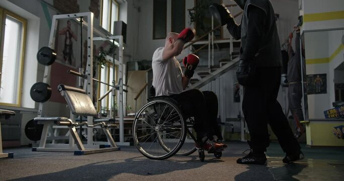 Training in the gym man smiling Paralympian with defective non-functioning legs sitting in a wheelchair Boxing with a coach in boxing gloves