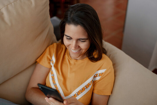 Young female text messaging on cellphone while lying down on couch in living room