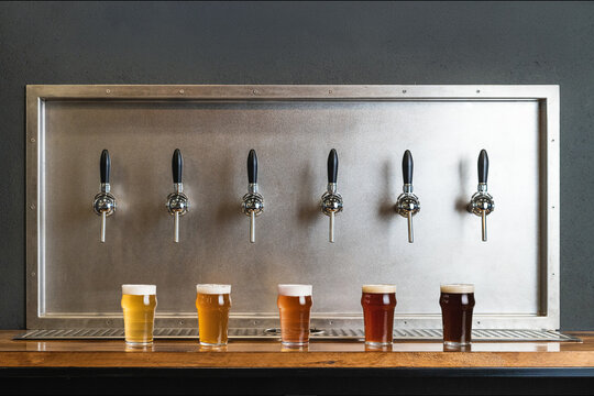 Different types of beer with foam in glass jugs against row of taps in bar on gray background