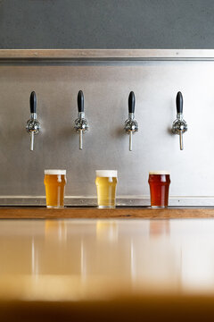 Different types of beer with foam in glass jugs against row of taps in bar on gray background