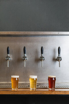 Different types of beer with foam in glass jugs against row of taps in bar on gray background