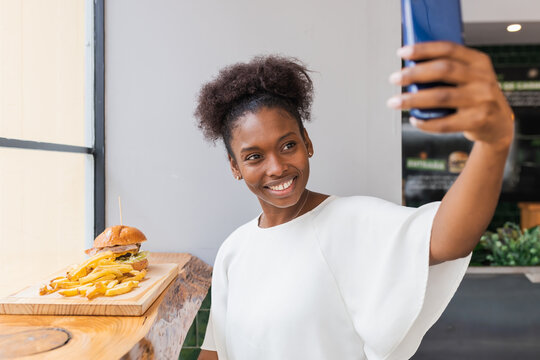 Side view joyful young African American female in white blouse enjoying delicious French fries and burger while taking selfie on smartphone in fast food restaurant
