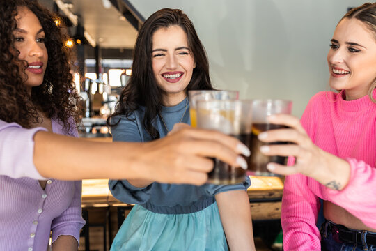 Joyful young multiethnic female best friends in stylish colorful outfits smiling and clinking glasses of refreshing juice and coke in modern bar