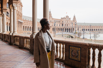 Young African American female in casual wear admiring views of grand palace with colonnade stucco work ornaments and columns while visiting Seville Spain