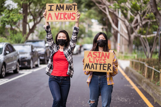 Ethnic females in masks holding posters protesting against racism in city street and looking at camera