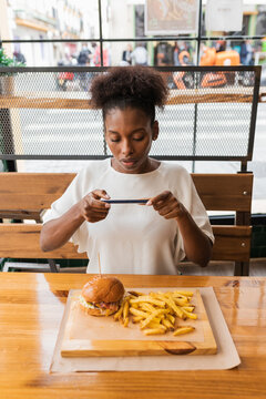 Front view of calm African American female in stylish wear taking pictures of yummy burger and fries served on high table in fast food restaurant.