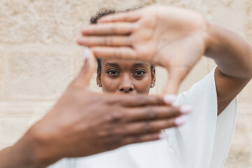 Happy young African American female in white blouse showing on camera framing sign with hands and looking at camera while standing against uneven wall