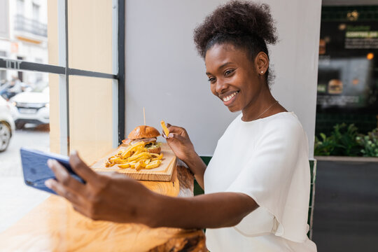Side view joyful young African American female in white blouse enjoying delicious French fries and burger while taking selfie on smartphone in fast food restaurant