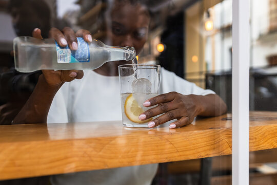 Through glass crop calm African American female in casual outfit pouring cold fresh water from bottle into glass with ice and lemon while sitting at high table in cafeteria