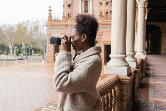 Side view young African American female photographer in warm coat taking pictures of historic city buildings on modern photo camera on early spring day