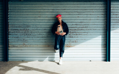 Transsexual man in trendy wear with purse and pink hair looking away against weathered wall on sunny day