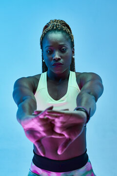 Young muscular African American female athlete in sportswear with clasped hands working out while looking at camera on blue background