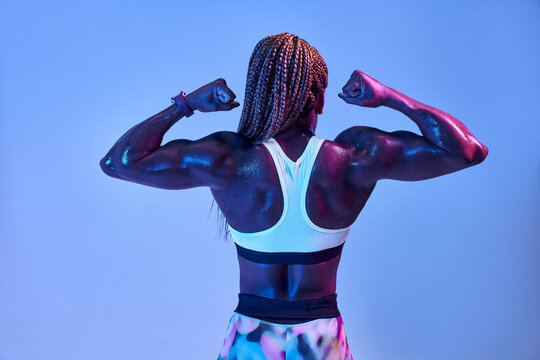 Back view of anonymous muscular African American female athlete with sweaty body showing biceps on blue background