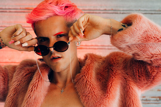 Young gay in stylish wear with tattoo, pink hair and manicure with fluffy jacket holding sunglasses while looking at camera
