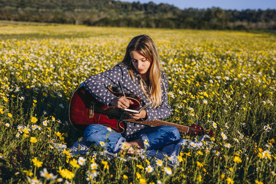 Young hipster woman sitting on a meadow in the countryside writing songs on notebook and playing guitar during summer sunlight.