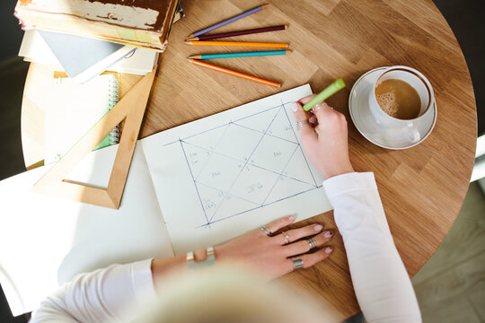 From above of crop unrecognizable female astrologist taking notes on paper with geometric drawing at desk with cup of coffee