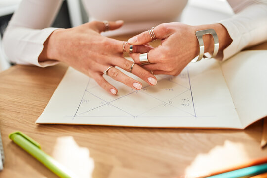 Crop unrecognizable female astrologist in rings and bracelets working at desk with paper album at home