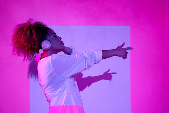 Side view young African American female listening to good music and pointing away while standing with eyes closed in neon lights