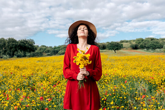 Female in hat with eyes closed holding blossoming yellow flowers in countryside field under cloudy sky