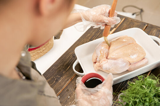 From above of crop anonymous female with pastry brush greasing raw poultry with soybean sauce while cooking at home
