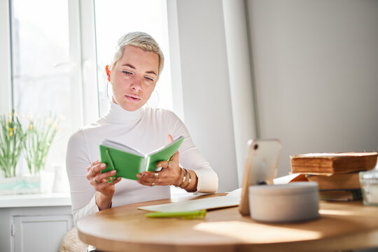Adult female astrologist reading notepad at desk with smartphone and books at home in sunlight
