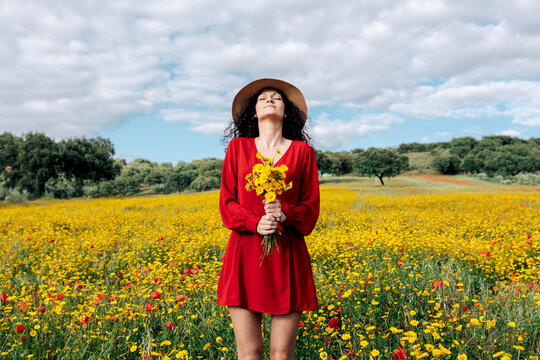 Female in hat with eyes closed holding blossoming yellow flowers in countryside field under cloudy sky