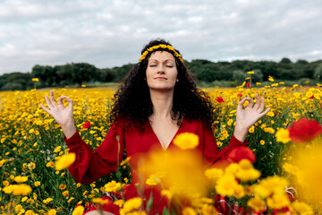 Concentrated female in flower wreath practicing yoga with closed eyes among blossoming daisies and Papaver on meadow in countryside