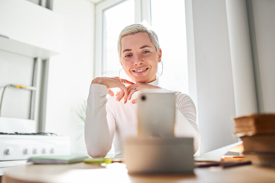 Smiling female blogger talking about astrology while recording video on cellphone at desk in sunlight