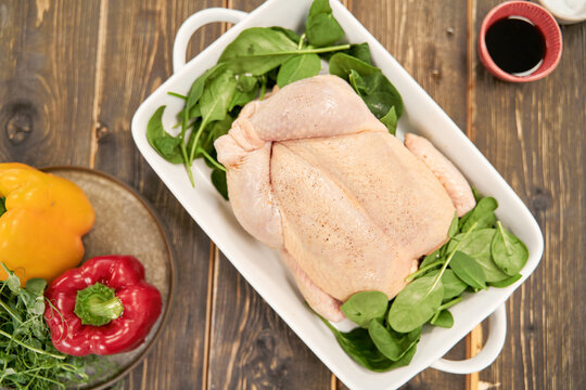Top view of whole uncooked poultry with fresh spinach foliage in baking dish between peppers and soy sauce on table