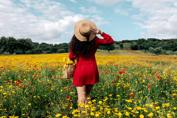 Back view anonymous trendy female in red sundress and handbag standing on blossoming field with yellow and red flowers and touching hat on warm summer day