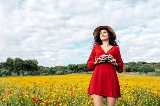 Smiling female in hat taking photo on vintage camera on meadow under cloudy sky