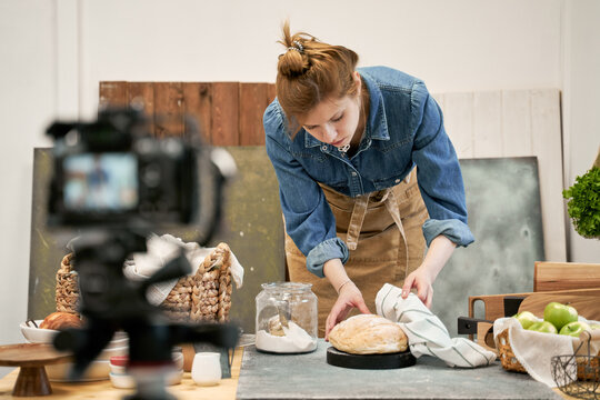 Young female blogger with fresh wheat bread against photo camera during cooking process at table in house