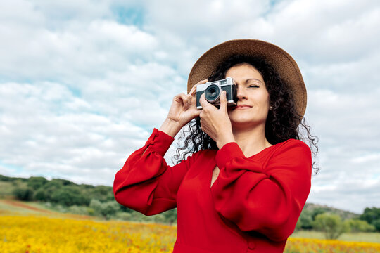 Smiling female in hat taking photo on vintage camera on meadow under cloudy sky