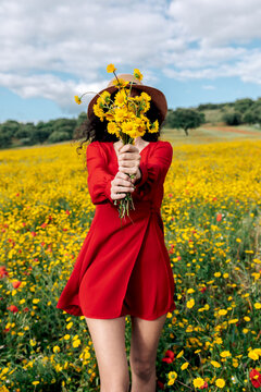 Anonymous female in hat covering face with blossoming yellow flowers in countryside field under cloudy sky