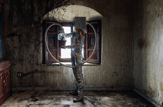 Side view unrecognizable person wearing silver costume with breathing apparatus and hose attached to potted plant standing in shabby abandoned room
