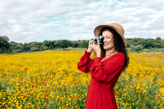 Smiling female in hat taking photo on vintage camera on meadow under cloudy sky