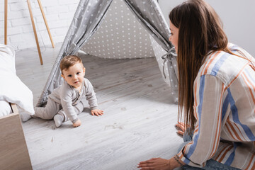 blurred mother looking at pleased baby son sitting near tipi in bedroom