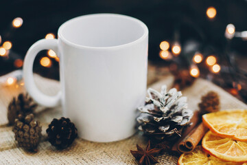 White empty cup and pine cones, anise, cinnamon, dried oranges on background of christmas lights. Winter time for hot cocoa and tea. Christmas holidays