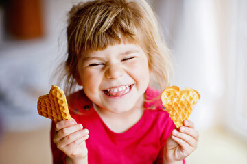 Portrait of happy little preschool girl holding fresh baked heart waffle. Smiling hungry toddler child with sweet biscuit wafer. Sweet sugar belgian waffles. - obrazy, fototapety, plakaty