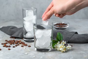 A woman's hand puts ice cubes in a transparent glass on a gray background.