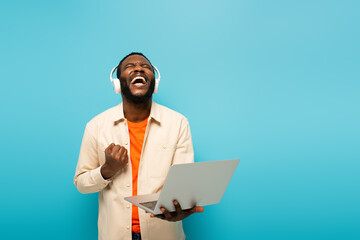 excited african american man in headphones showing success gesture while standing with laptop isolated on blue