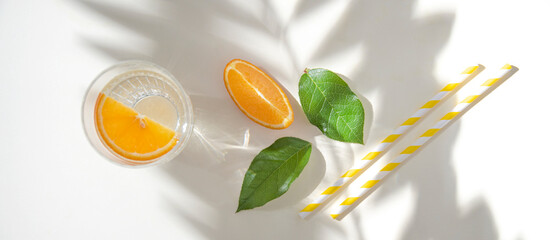 A glass of mineral water and a slice of orange on a light  table with fresh juicy fruit oranges and green leaves with hard shadows. Summer time. Top view
