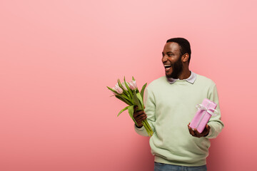 laughing african american man with gift box and tulips looking away on pink background